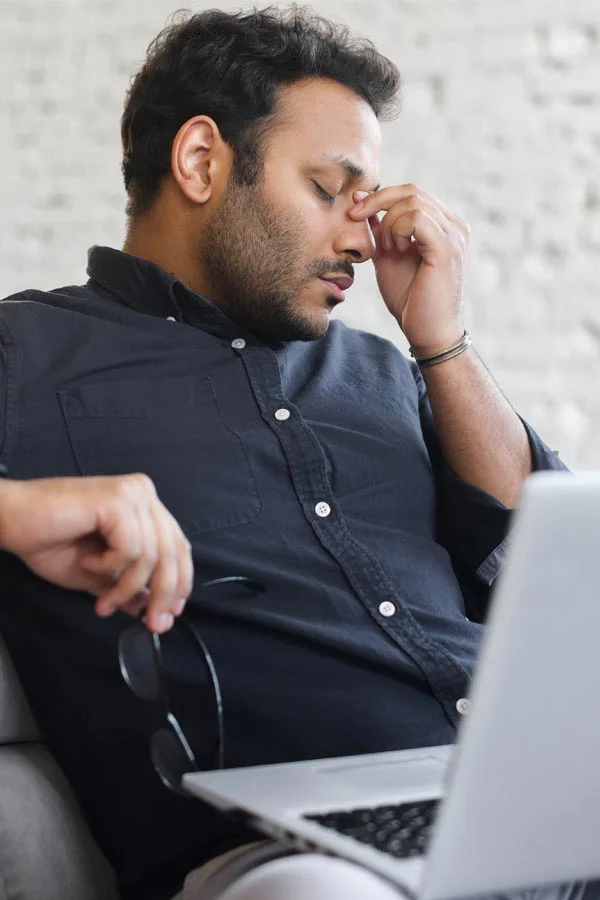 Adrenal-Fatigue-Treatment A middle-aged man with a laptop closing his eyes and rubbing his nose. He needs treatment for Adrenal Fatigue Syndrome from Julie Kane, NP of Bird Island Wellness in Calgary.