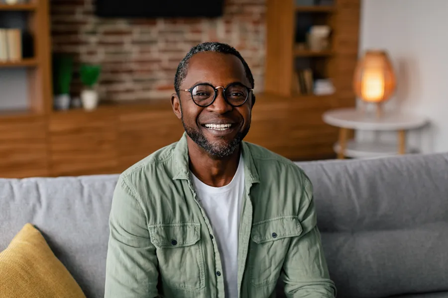 Andropause-Doctor A middle aged black man in a green button up shirt sits on his couch smiling, happy with his treatment for andropause and low testosterone from Julie Kane, NP of Bird Island Wellness in Calgary.