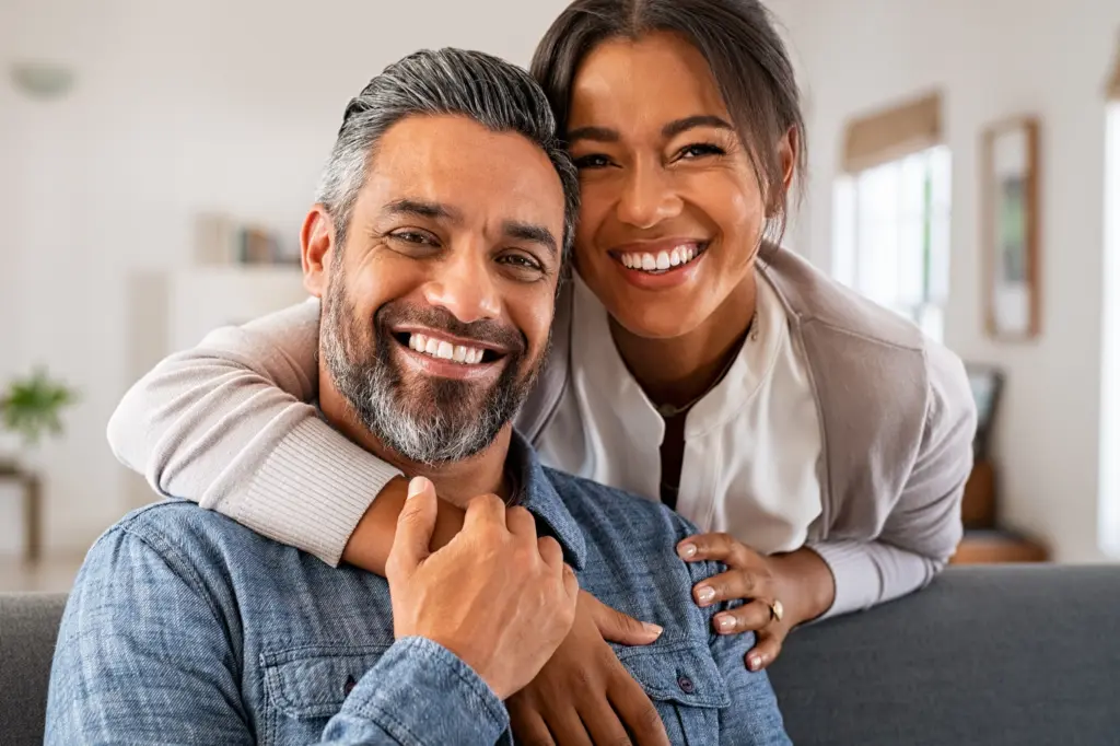 A middle-aged couple embrace in their living room smiling at the camera. Schedule Bioidentical Hormone Replacement Therapy from Julie Kane, NP of Bird Island Wellness in Calgary.