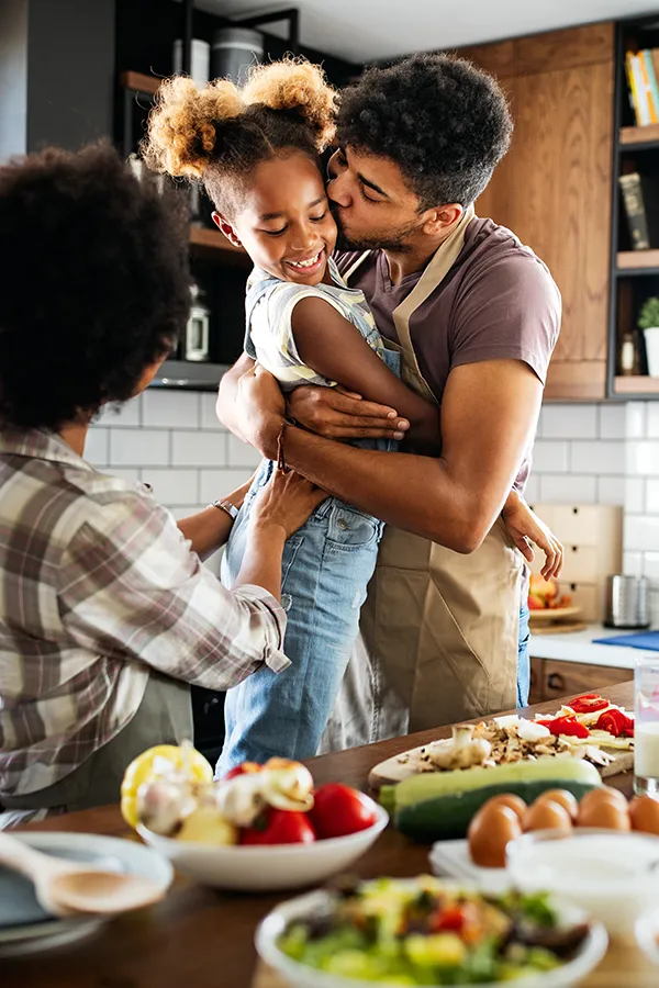 Gut-Health-Treatment A man holds his daughter and kisses her cheek as the family makes a healthy dinner in the kitchen. Get help for digestive issues and gut health from Julie Kane, NP of Bird Island Wellness in Calgary.