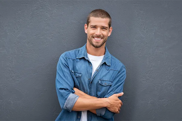 Hormones-for-Men-Doctor A man in a denim shirt stands smiling against a gray-blue wall, pleased with his testosterone hormone therapy from Julie Kane, NP of Bird Island Wellness in Calgary.