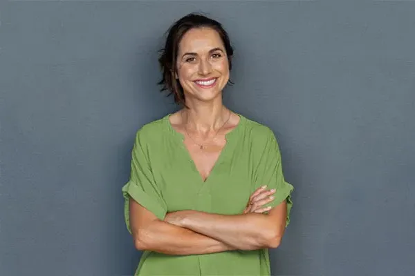 Hormones-for-Women-Doctor A woman in a green shirt stands smiling against a gray-blue wall, pleased with her hormone therapy from Julie Kane, NP of Bird Island Wellness in Calgary.