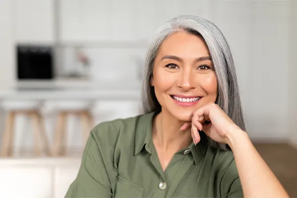Menopause-Clinic An asian woman with long gray hair and a green shirt sitting and smiling on her couch, happy with her menopause care from [NAME} in Calgary.