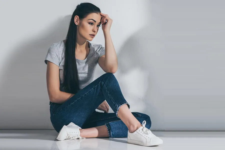 PCOS-Doctor A young brunette woman in a gray t-shirt and jeans sits against a gray wall with her head resting on her hand and a distressed expression. Get help for Polycystic Ovarian Syndrome (PCOS) from Julie Kane, NP of Bird Island Wellness in Calgary.