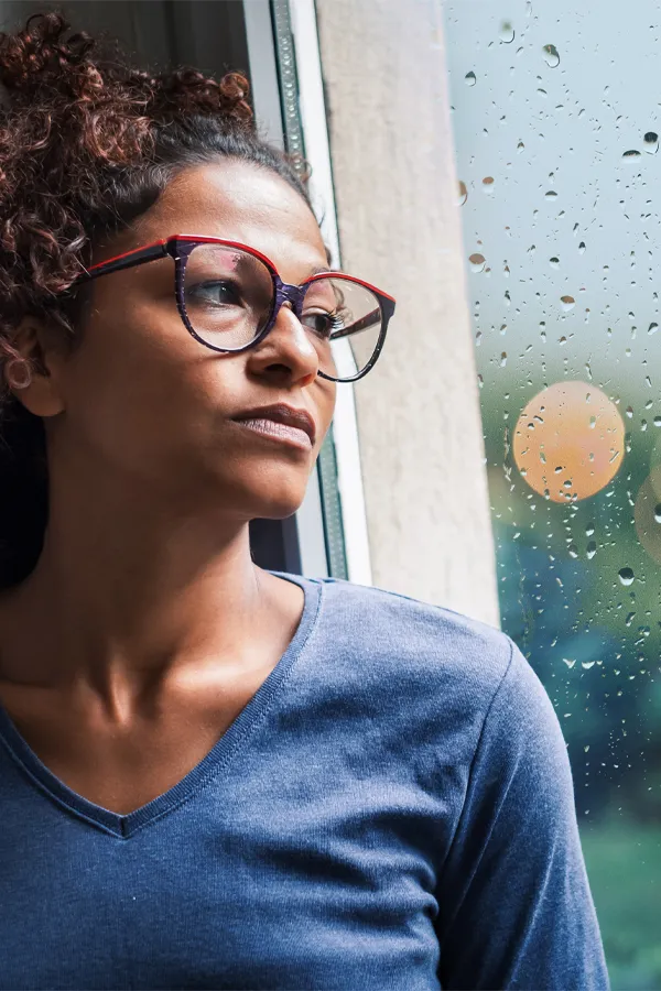 PMDD-Treatment A black woman with with a somber expression and glasses and a gray shirt stares out the window on a rainy day. Get treatment for Premenstrual Dysphoric Disorder from Julie Kane, NP of Bird Island Wellness in Calgary.