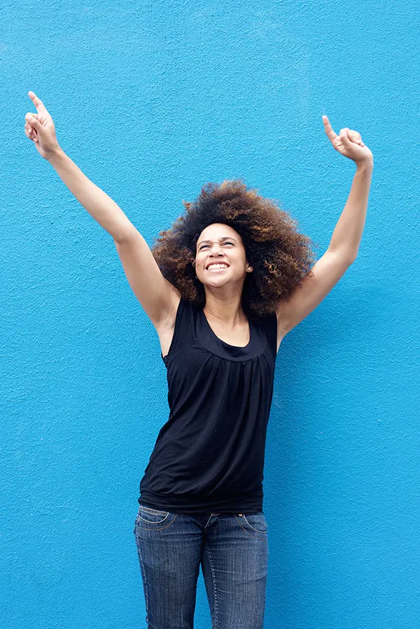 PMS-Treatment A woman in a dark blue tank top standing in front of a bright blue wall, raising her arms in celebration of relief from PMS from Julie Kane, NP of Bird Island Wellness in Calgary.
