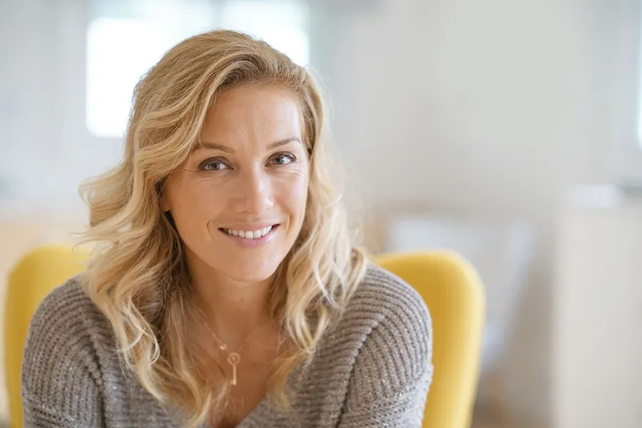 Perimenopause-Doctor A blonde woman wearing a a gray sweater sits inside in a yellow chair, smiling and happy with her perimenopause treatment from Julie Kane, NP of Bird Island Wellness in Calgary.
